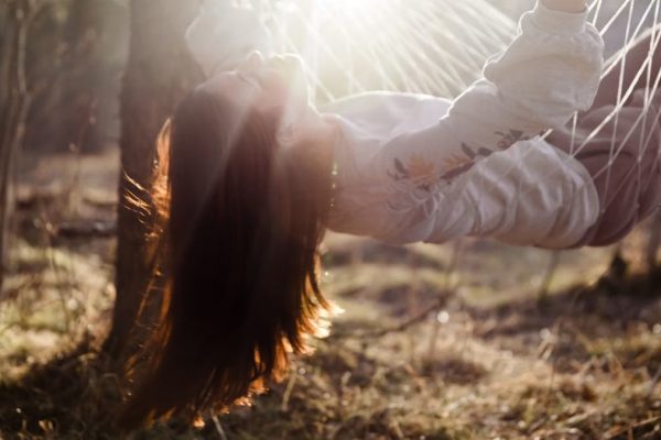Young woman with long hair lies playfully in a sunlit hammock outdoors, embracing nature and sunlight.